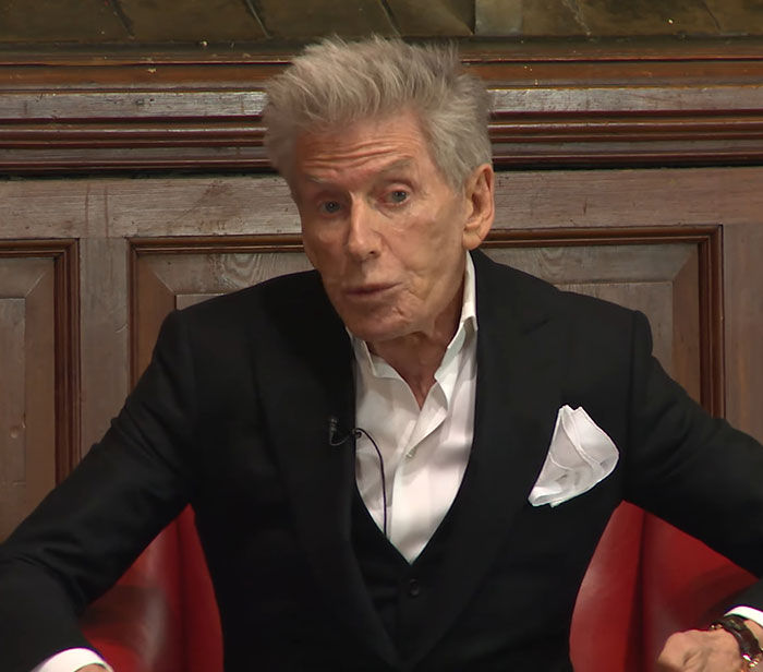 Calvin Klein, 83, wearing a black suit and white shirt, seated and speaking with a serious expression indoors.