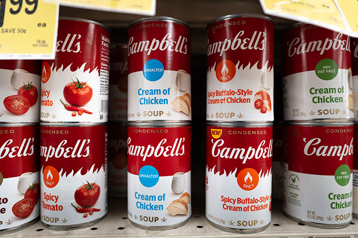 Cans of Campbell's soup including Cream of Chicken and Spicy Buffalo-Style flavors on a supermarket shelf.