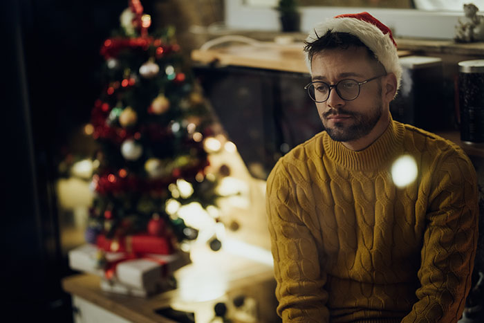 Man wearing Santa hat and glasses, sitting by a decorated Christmas tree, appearing sad about Christmas affordability.