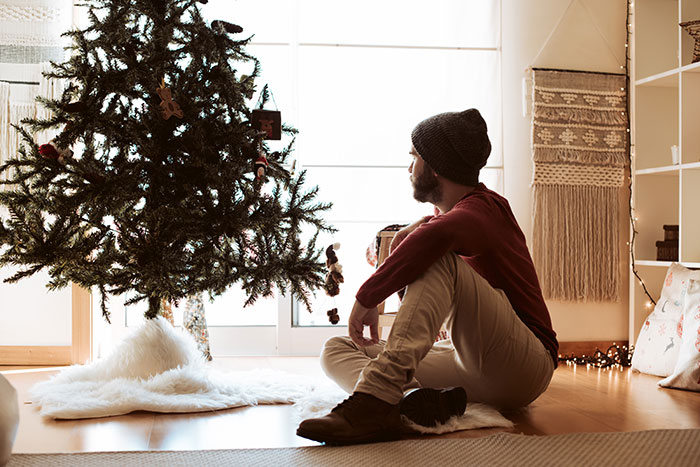 Man sitting on floor by Christmas tree, looking thoughtful, reflecting on Christmas affordability struggles.