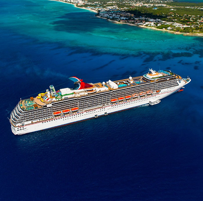 A large Carnival cruise ship sailing near a tropical coastline with clear blue water under bright sunlight.