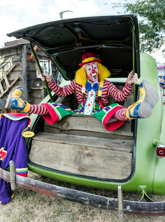 Clown with colorful outfit sitting in the open trunk of a green vintage car, eerie scene truckers have seen on the road.