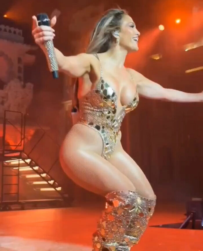 Jennifer Lopez performing in a risqué gold outfit on stage during a billionaire's wedding event with dramatic lighting. Jennifer Lopez performing in a risqué gold outfit on stage during a billionaire's wedding event with dramatic lighting.