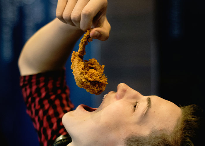 Person in a red and black shirt holding fried chicken above their open mouth in a chaotic food moment witnessed by many.