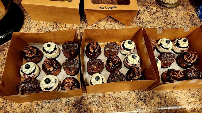 Three boxes of assorted cupcakes with chocolate, cream, blueberries, and drizzled toppings on a kitchen counter surface.