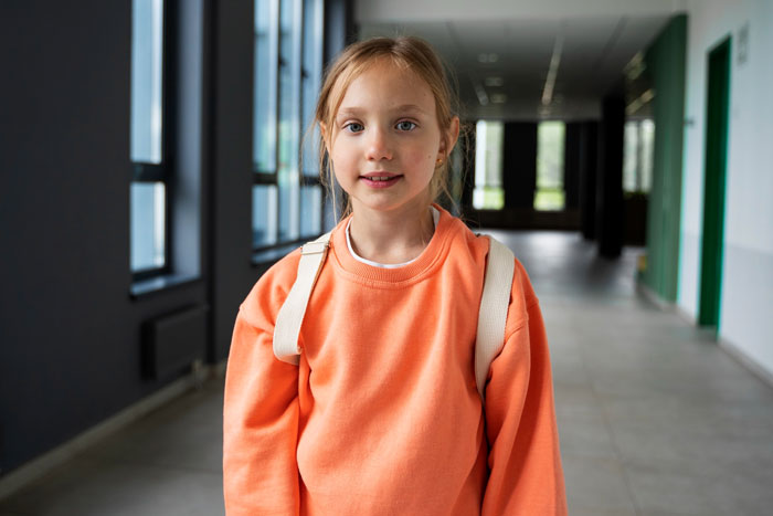 Young girl in orange sweatshirt with backpack in school hallway, representing daughter affected by native language ban conflict.