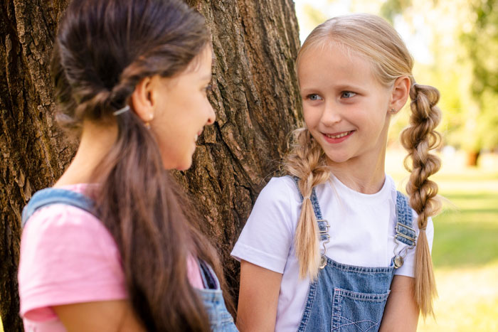 Two young girls smiling and talking near a tree, highlighting a daughter's use of her mom&rsquo;s native language banned by dad.
