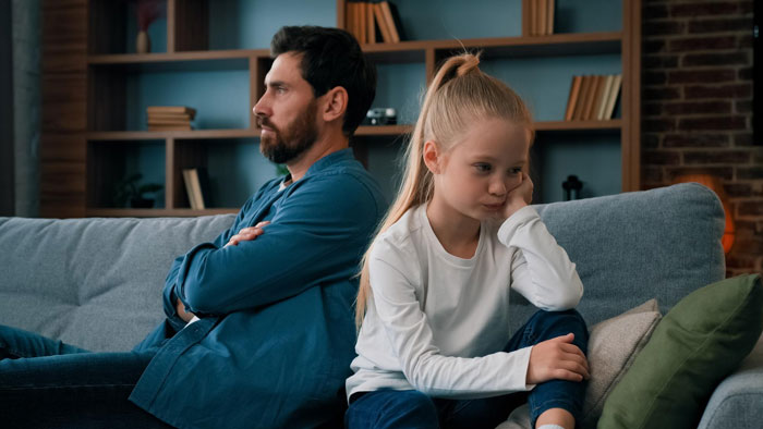Father and daughter sitting apart on a couch looking upset, illustrating conflict over mom&rsquo;s native language ban.