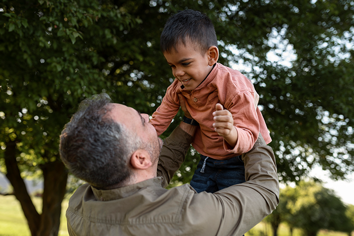Man lifting young boy outdoors under trees, symbolizing family health fund dispute, woman suing half-brother over plastic surgeries.