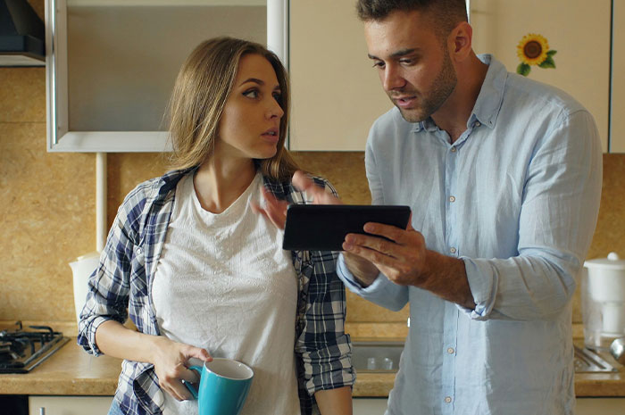 A couple discussing finances in a kitchen while looking at a tablet, highlighting choosing nose job over kids college funds.