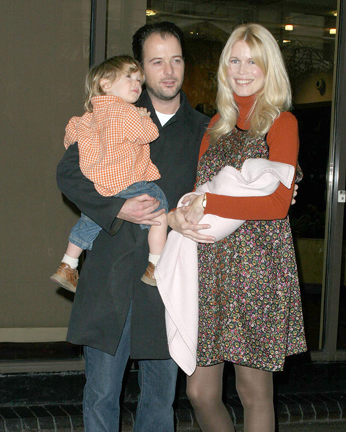 Claudia Schiffer&rsquo;s lookalike daughter in a floral dress holding a baby, standing with a man holding a toddler outside a building