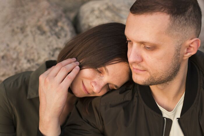 Young couple sitting outdoors, woman resting head on man’s shoulder, expressing emotions related to regret having kids.