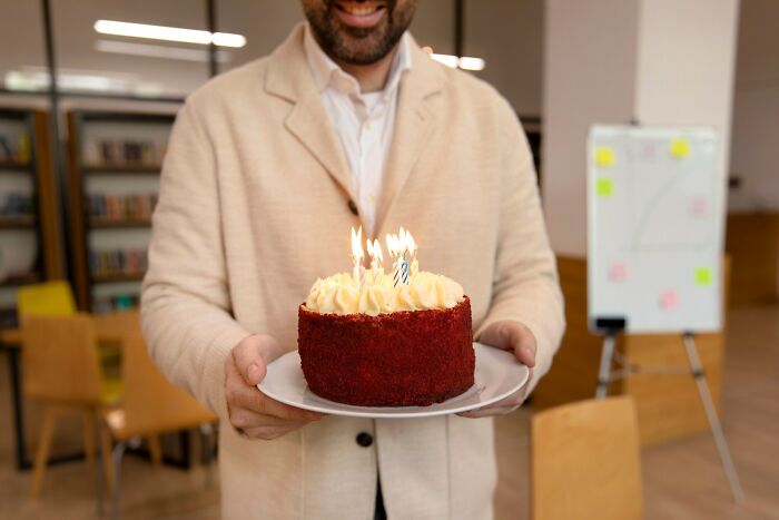 Man holding a small cake with lit candles in an office setting, representing infuriating examples experienced at work.