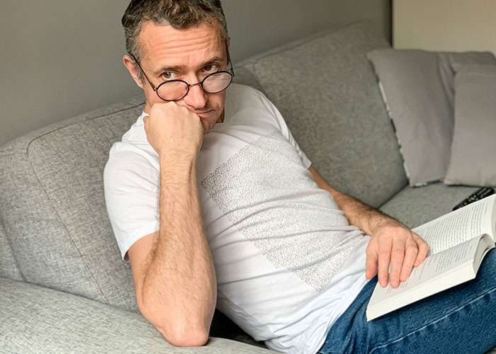 Man wearing glasses sitting thoughtfully on a gray couch reading a book, reflecting on life in the USA or Europe debate.