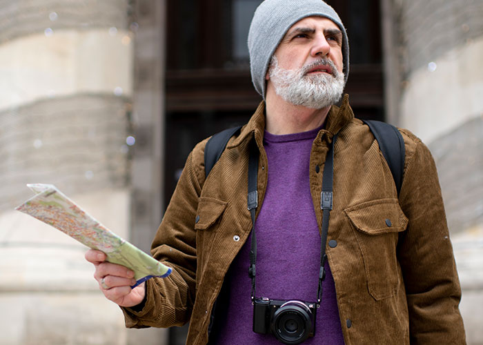 Middle-aged traveler with gray beard holding a map and camera, exploring life in the USA and Europe debate.