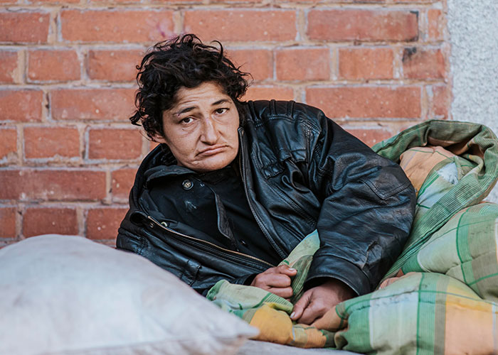 Person resting outdoors on a blanket against a brick wall, illustrating life challenges in the USA versus Europe debate.