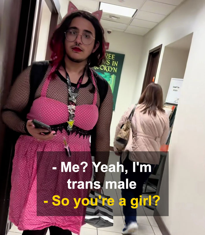 Bearded trans man in pink dress standing in hallway with a woman walking away in a restroom debate setting.