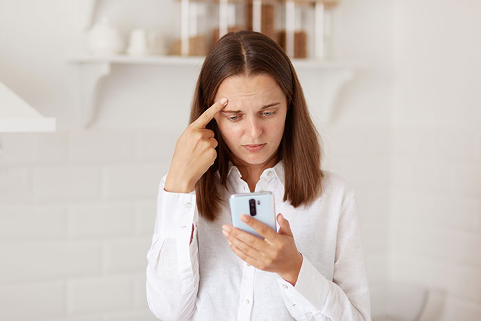 Woman in white shirt looking confused and concerned while checking phone, reflecting shock over half-sis medical bills lie.