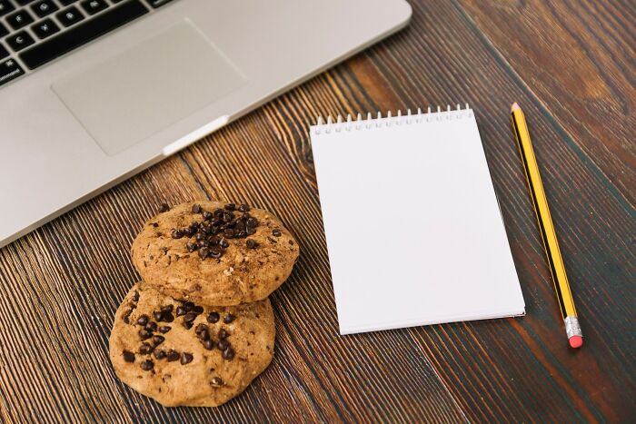 Laptop keyboard, two chocolate chip cookies, blank notepad, and pencil on a wooden desk illustrating compulsory things in life.