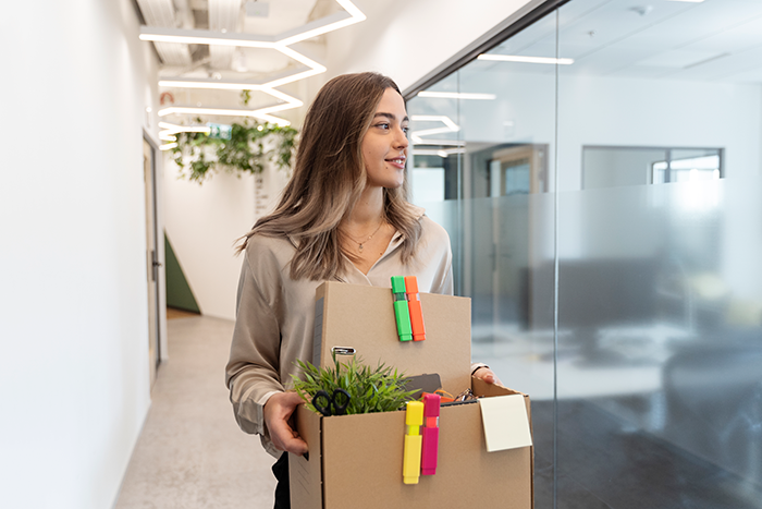 Young employee carrying a box of office items during an office swap, while leaving a modern workplace hallway.