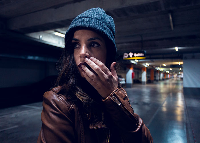 Woman in a brown leather jacket and blue beanie looking anxious in a dimly lit parking garage, evoking kidnapping fear.