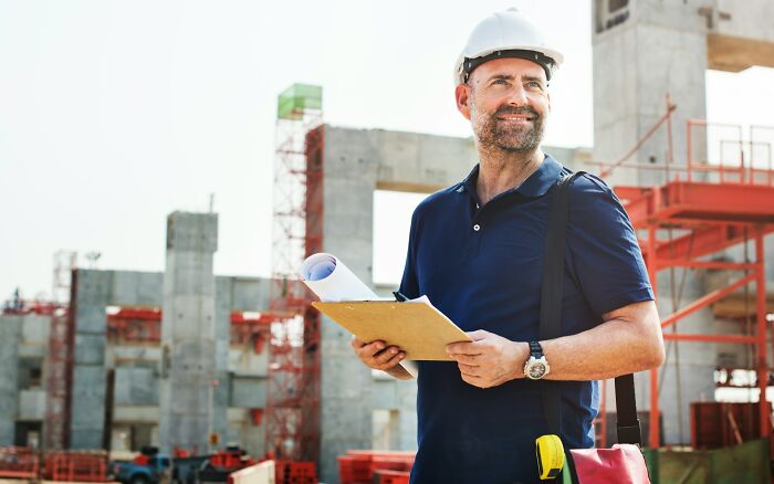 Construction worker with helmet holding blueprints at site, illustrating work secrets about trades like plumbers and baristas.