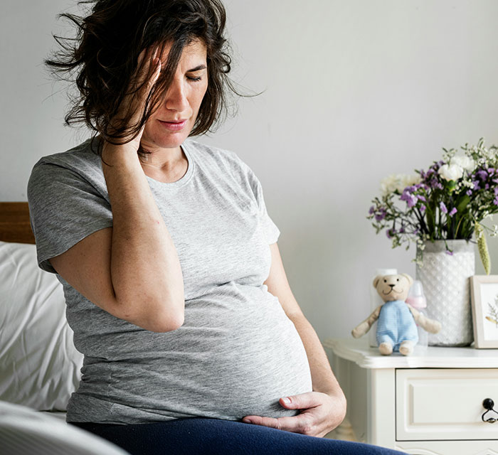 Pregnant woman sitting on bed holding her belly and head, showing a need for husband respect and support.