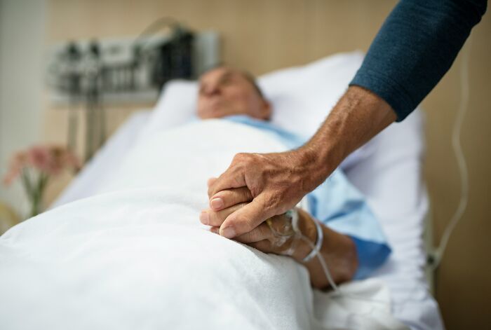 Patient lying in hospital bed holding hands with a visitor, capturing eerie moments patients said that still haunt doctors.