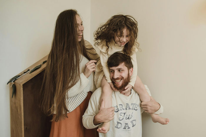 Happy young father with child on shoulders and woman smiling indoors, symbolizing dad busted stealing son&rsquo;s inheritance.