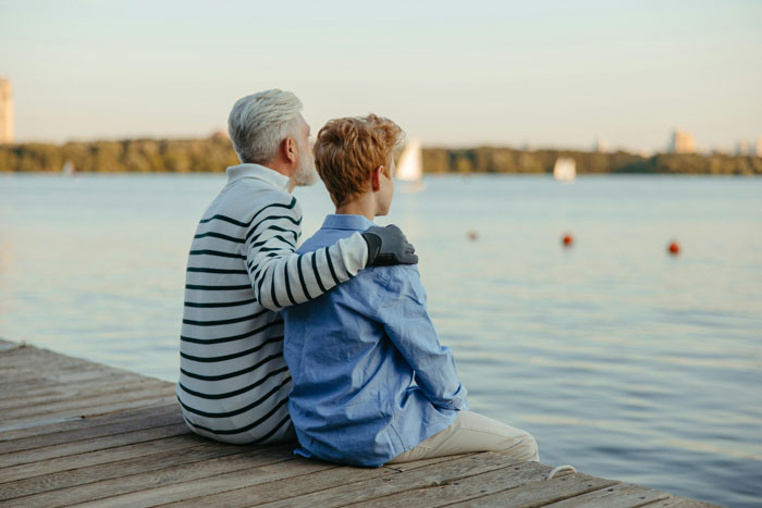 Older man with gray hair and teen boy sitting on dock by lake, representing family and inheritance themes.