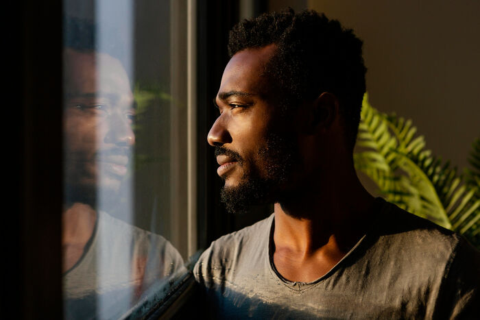 Man with beard looking thoughtfully out window, reflecting on dating older people stories while sitting indoors near plant.