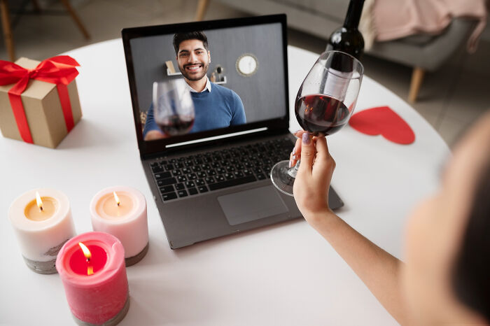 Person holding wine glass during a virtual date on laptop, surrounded by candles and a gift for dating older people stories single.