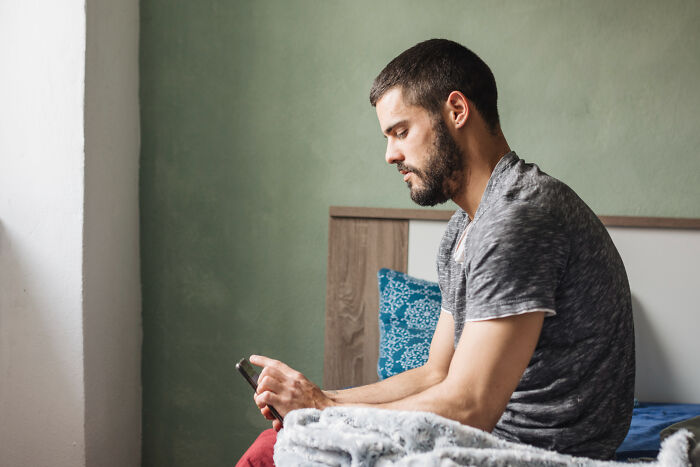 Man sitting on bed using smartphone, reflecting on dating older people stories single in a quiet bedroom.