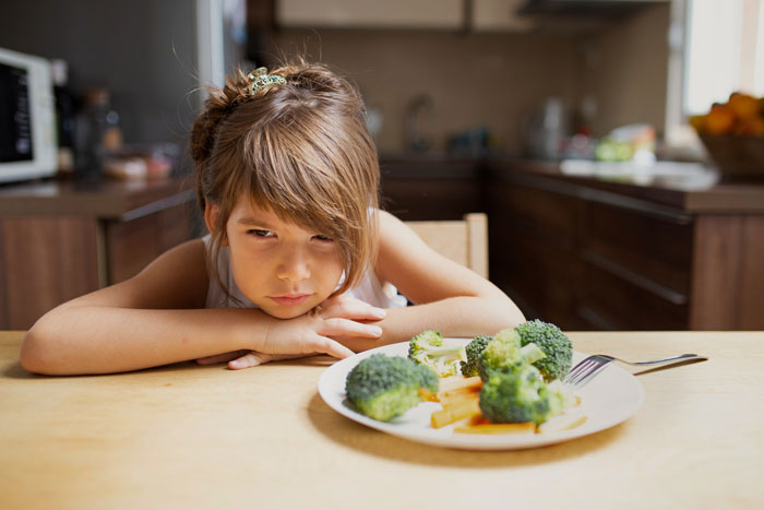 Young girl looking unhappy at a plate of broccoli and fries in a kitchen, reflecting foodie woman struggles with future meals. Young girl looking unhappy at a plate of broccoli and fries in a kitchen, reflecting foodie woman struggles with future meals.