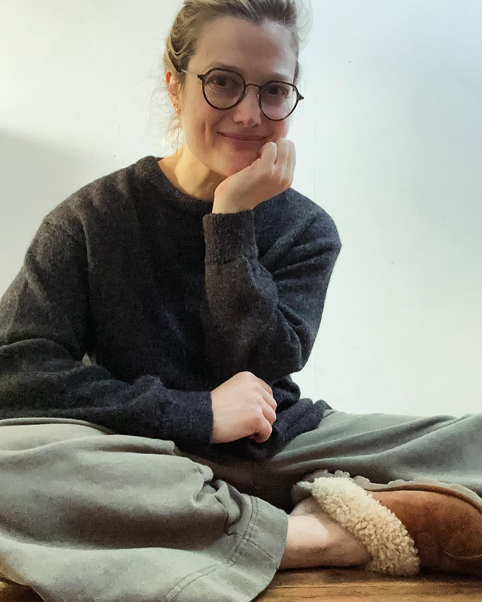 Alison Sudol sitting casually indoors wearing glasses, a dark sweater, and cozy slippers, smiling gently at the camera