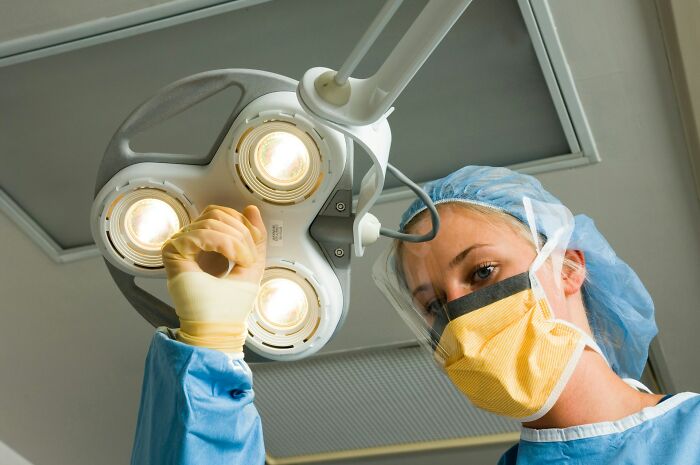 Dentist adjusting overhead dental light, wearing protective mask, gloves, and surgical cap in a clinical setting.