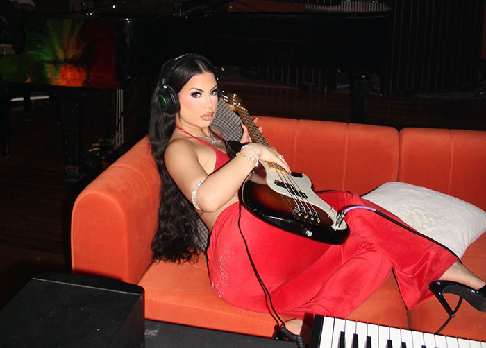 Singer DELAROSA wearing headphones and red outfit, holding a bass guitar while reclining on an orange couch. Singer DELAROSA wearing headphones and red outfit, holding a bass guitar while reclining on an orange couch.