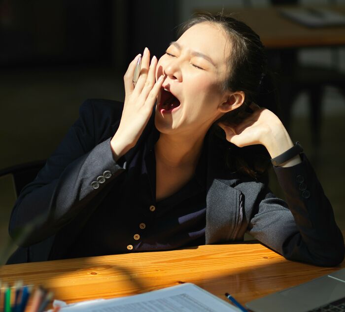 Young woman yawning at a wooden desk, appearing tired and stressed about plans to move for DIL&rsquo;s job conflict.