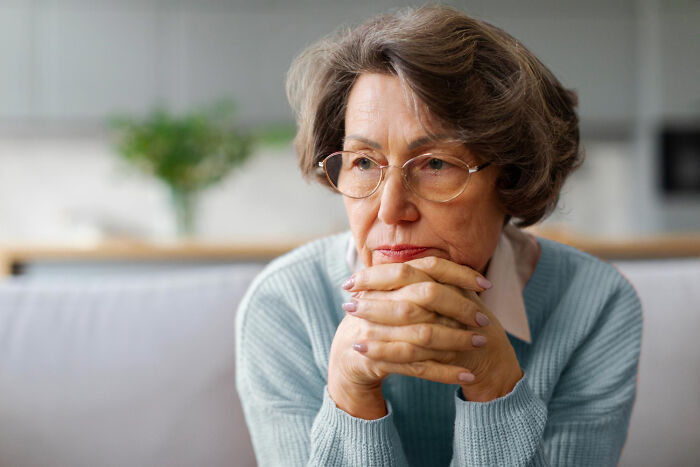 Older woman wearing glasses and a blue sweater looking thoughtful and concerned while sitting indoors at home.
