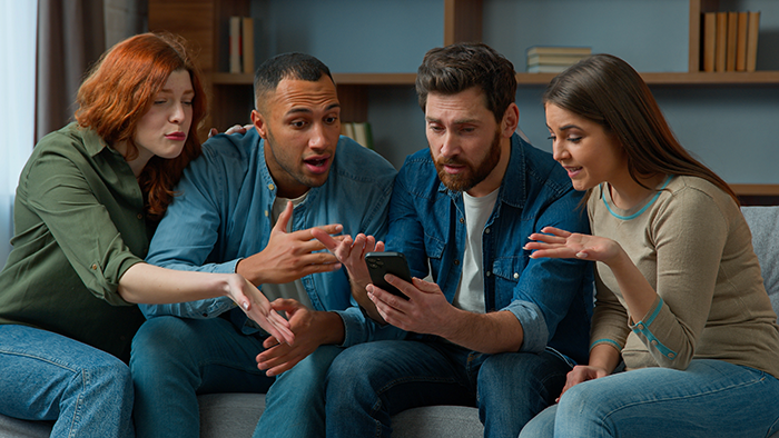 Four young adults sitting on a couch, looking shocked while viewing a phone, illustrating family health fund dispute.