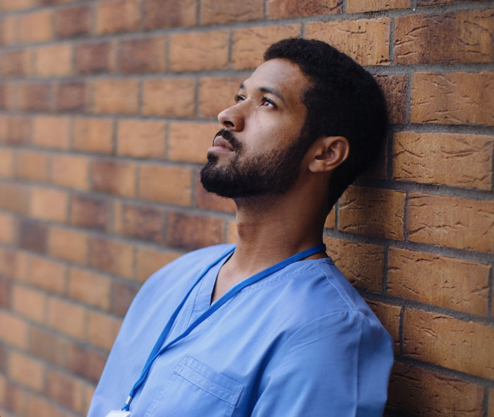Young surgeon in blue scrubs leaning against a brick wall, reflecting thoughtfully on medical mistakes and challenges.
