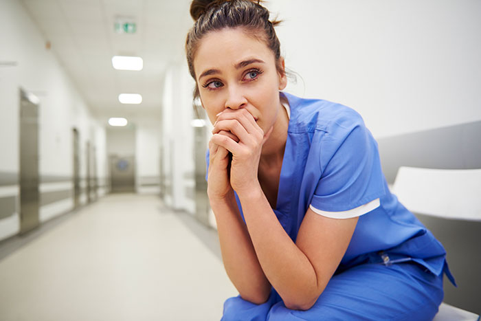 Worried female doctor in blue scrubs sitting in hospital corridor reflecting on medical mistakes and surgeon challenges.