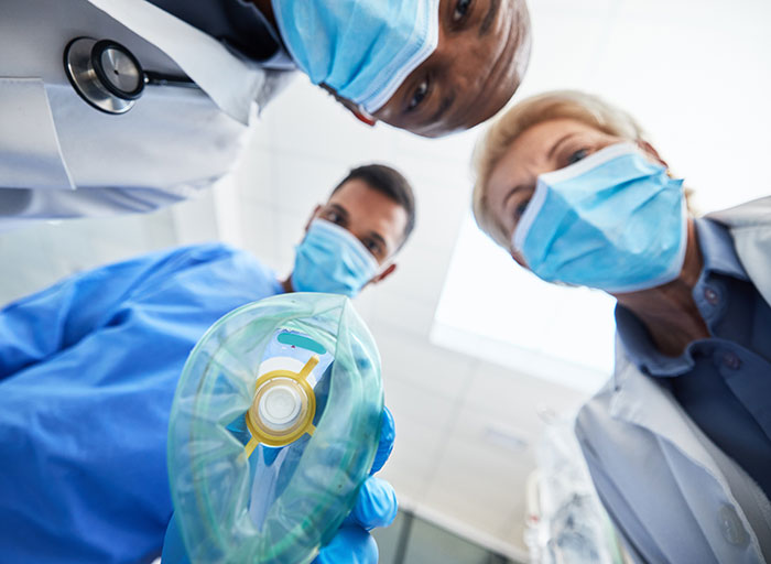 Surgeons and doctors wearing masks look down while preparing to use an anesthesia mask in a medical setting.
