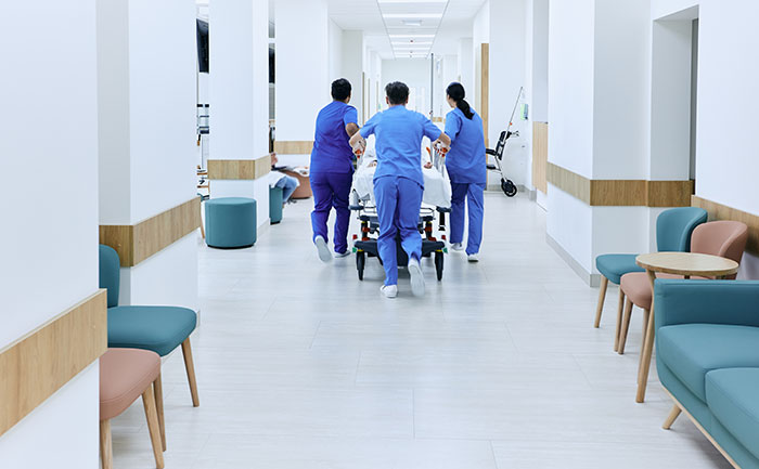 Three surgeons and doctors in blue scrubs rush a patient on a stretcher down a hospital corridor.