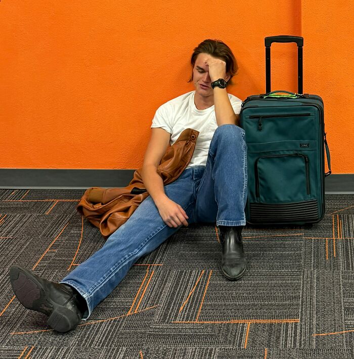 Young man sitting on floor by suitcase and orange wall, appearing contemplative, representing people who disappeared to start new lives.