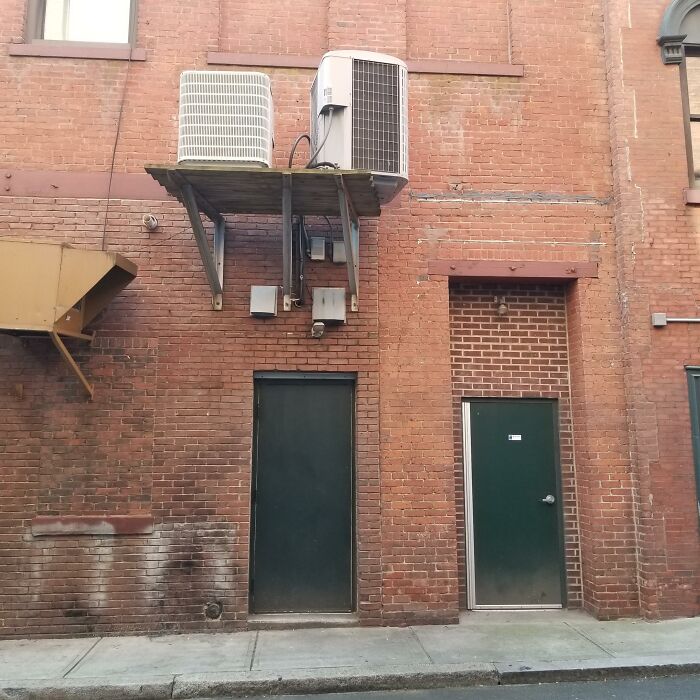Two adjacent dark green doors on a brick wall, with air conditioning units awkwardly installed above one, showing lack of effort.