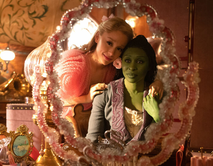 Cynthia Erivo and Ariana Grande reflected in a decorative mirror during a bizarre interaction scene.