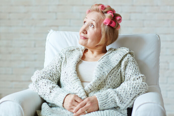Elderly woman with pink hair rollers sitting in a chair, wearing a cozy sweater, heart racing moment concept.