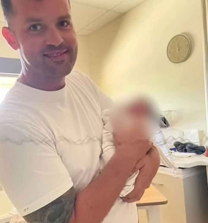 Man with tattooed arm holding a blurred toddler indoors, related to teen unsurprised dad took his life after leaving 2YO in hot car.
