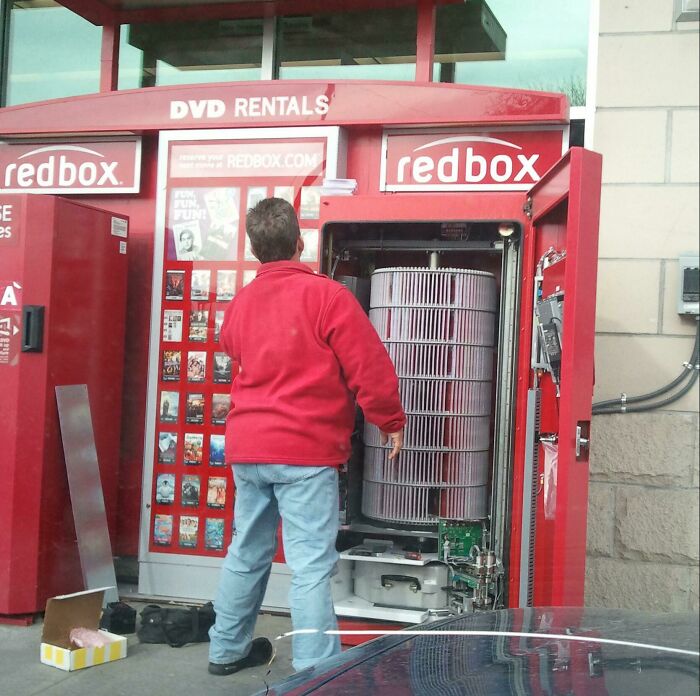 Man in red jacket repairing the insides of a Redbox DVD rental machine showing internal mechanical components.